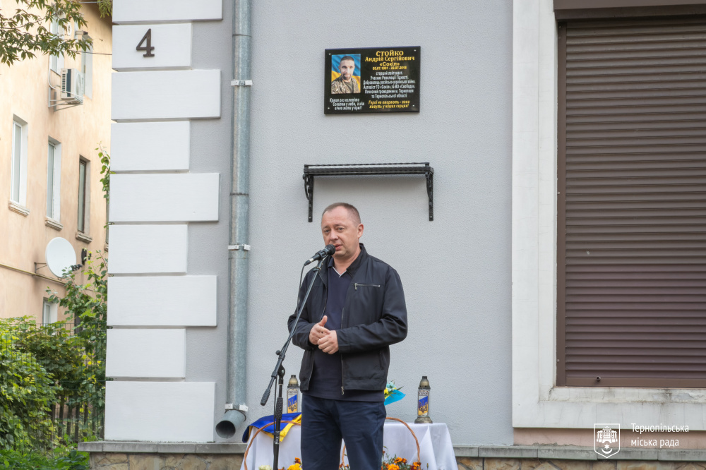 Чоловік виступає біля меморіальної дошки, стоячи перед мікрофоном. Позаду — стіл із квітами та лампадками.