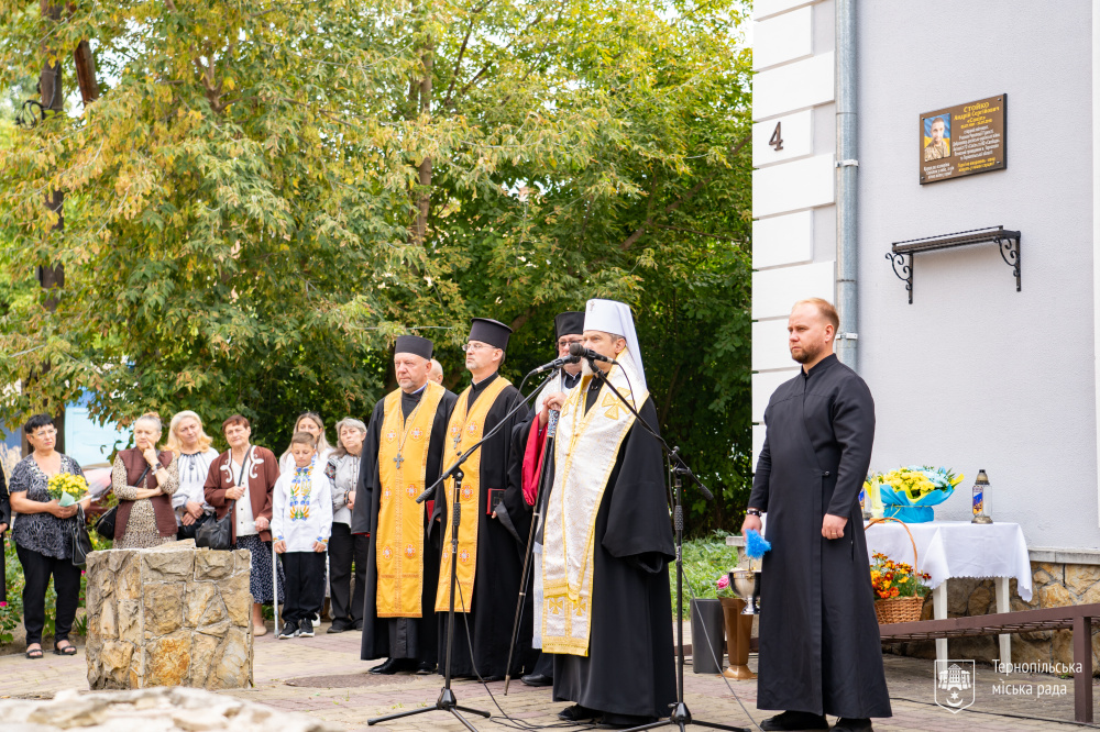 Священнослужителі стоять біля мікрофонів під час пам’ятного заходу. Праворуч — чоловік у чорному одязі, позаду присутні люди.