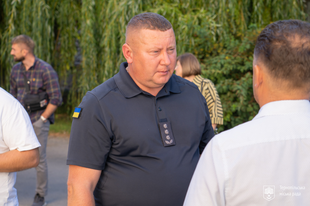 На фото працівник поліції у темній формі розмовляє з чоловіком у світлій сорочці. На рукаві поліцейського видно прапор України. На задньому плані — інші учасники навчання