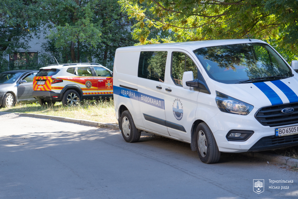 На фото припарковані службові автомобілі. На передньому плані — білий автомобіль аварійної служби водоканалу. Позаду нього — автомобіль ДСНС із позначенням екстрено-рятувальної служби.