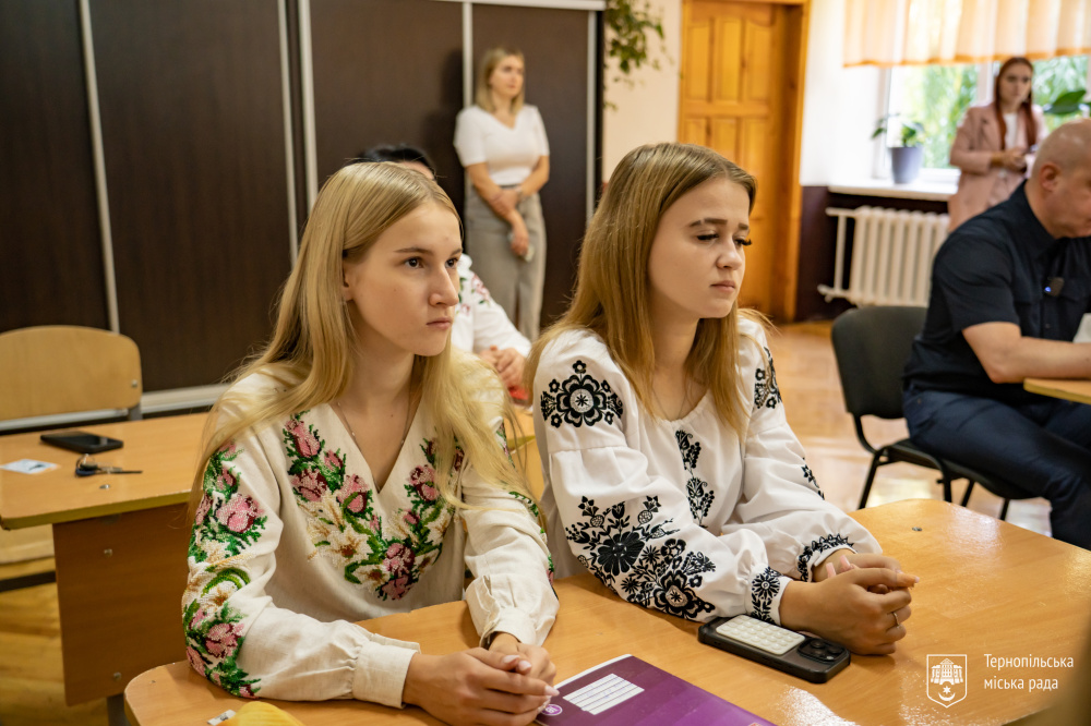ві учениці сидять за партою та слухають урок. На них вишиті сорочки.