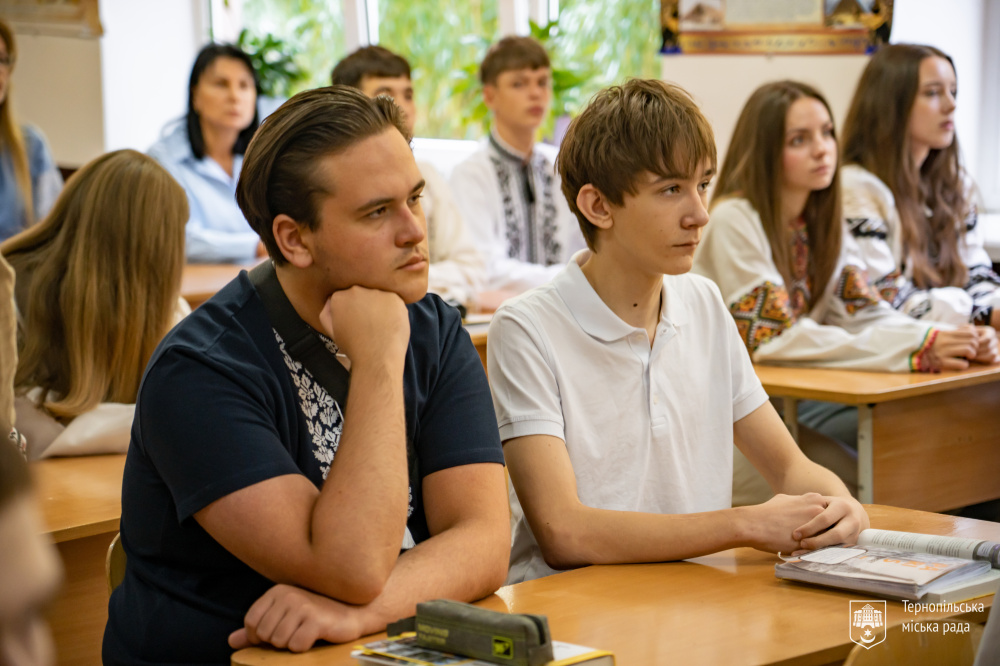 Учні сидять за партами та слухають урок. На передньому плані — хлопець у темній сорочці та хлопець у білій футболці.