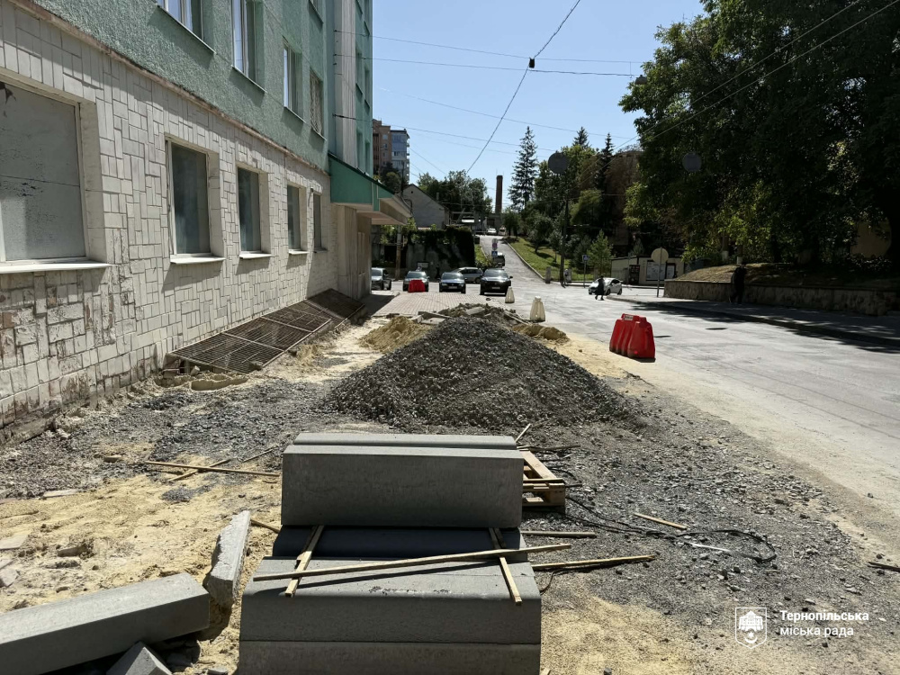 Вздовж довгої світло-зеленої будівлі ділянку тротуару засипано гравієм, а на передньому плані чекають на монтаж нові бетонні бордюри.