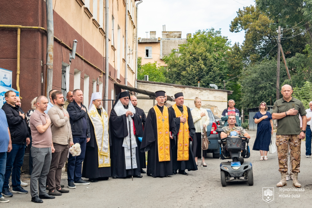 Група людей, священнослужителі та військовий стоять біля будинку під час пам’ятного заходу.
